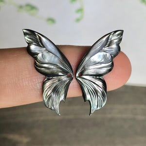 Puede incluir: Dos colgantes iridiscentes con forma de alas de mariposa. Las alas son de un gris oscuro nacarado con vetas intrincadas y un brillo sutil. Cada ala tiene un pequeño agujero cerca del centro. Las alas están sujetas por el dedo de una persona.