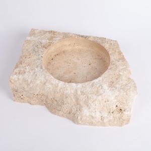 May include: A natural stone bowl with a rough, textured exterior and a smooth, circular interior. The bowl is made of travertine and has a light beige color.