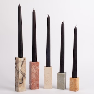 May include: Five black taper candles of varying heights, each set in a rectangular stone holder. The holders are in different colours and textures, including beige, red, and grey, against a white background.