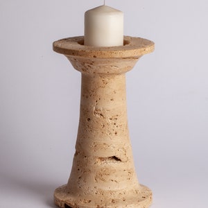 Könnte beinhalten: Ein beigefarbener Kerzenhalter aus Stein mit einer weißen Kerze darauf. Der Halter hat eine breite Basis und einen hohen, schlanken Stiel.