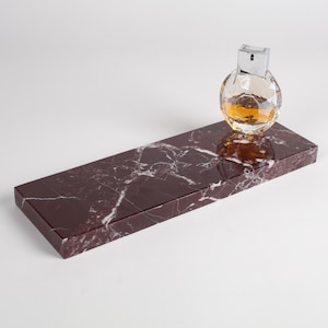 May include: A rectangular dark maroon marble tray with white veining patterns. A clear, spherical glass perfume bottle with a silver cap and amber-colored liquid sits on the right side of the polished stone surface.