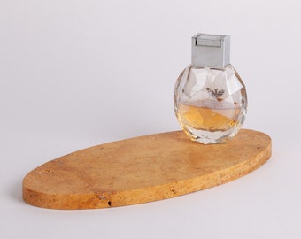 Puede incluir: Una botella de perfume de vidrio transparente con una tapa plateada se encuentra sobre una bandeja de corcho ovalada de color marrón claro. La botella tiene un diseño facetado y contiene un líquido amarillo claro.