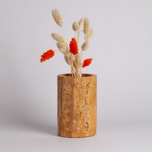 May include: A cylindrical vase made of travertine stone with a natural, textured finish. The vase is filled with dried flowers, including orange and white blooms.