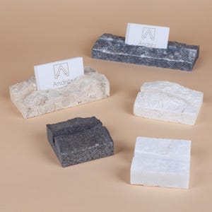 May include: Several stone business card holders in different colours and textures. Each holder displays a white business card with the name "Andrés" and a logo. The holders are in shades of grey, white, and beige.