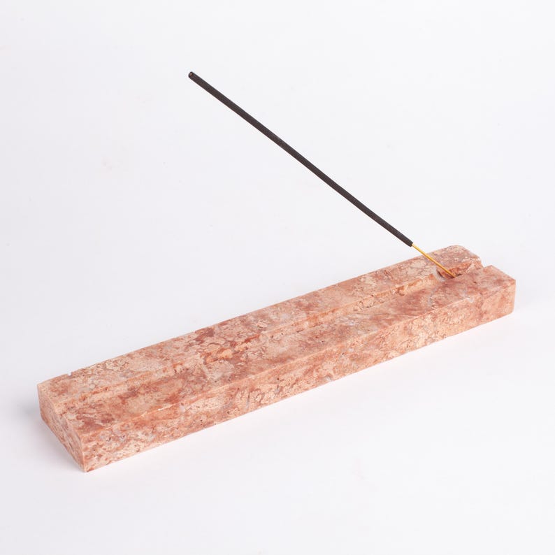 Pode incluir: Um porta-incenso retangular feito de m&aacute;rmore castanho-avermelhado. O suporte tem duas ranhuras para segurar varetas de incenso. Uma vareta de incenso preta com ponta dourada &eacute; colocada em uma das ranhuras.