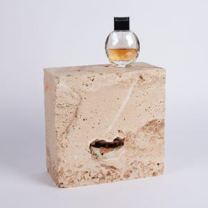 May include: A clear glass bottle of perfume with a black cap sits on top of a beige travertine stone block.