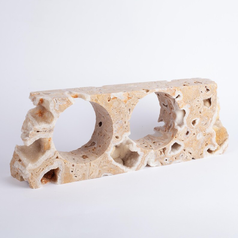 May include: A beige stone shelf with two circular openings. The shelf has a rough, natural texture and is made of a light-colored stone.