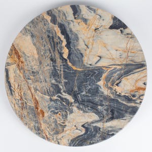 Pode incluir: Um tampo de mesa circular em pedra com uma superfície polida que apresenta um padrão complexo de veios em espiral azul-marinho escuro, creme e branco, acentuado por estrias proeminentes de cor laranja ferrugem e castanho dourado.