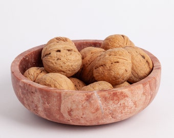 Puede incluir: Un cuenco de mármol rosa lleno de nueces. El cuenco tiene un aspecto natural y rústico con vetas y remolinos visibles.
