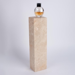 Può includere: Un flacone di profumo in vetro trasparente con tappo nero poggia su un piedistallo in travertino beige.