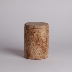 May include: A cylindrical stone container with a lid. The container is made of travertine and has a natural, earthy brown color with brown and white veining.
