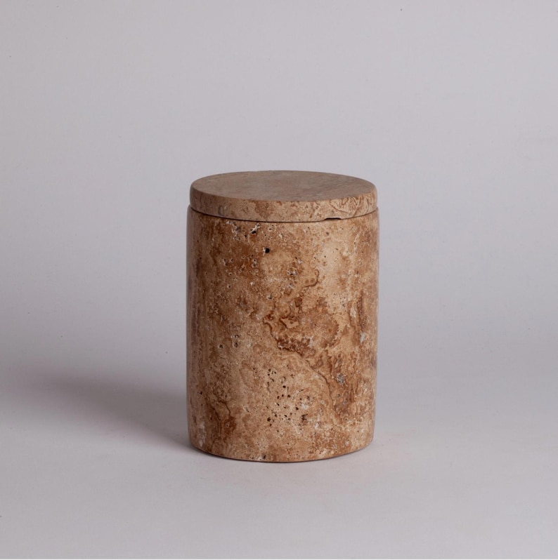 May include: A cylindrical stone container with a lid. The container is made of travertine and has a natural, earthy brown color with brown and white veining.