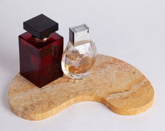 Puede incluir: Dos botellas de perfume sobre una bandeja de piedra de color marrón claro. La botella de la izquierda es una botella de vidrio rojo oscuro con una tapa negra. La botella de la derecha es una botella de vidrio transparente con una tapa plateada y un diseño facetado.
