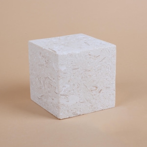 May include: A white cube-shaped pedestal made of a textured material, resembling marble or stone. The pedestal is a neutral color and has a smooth surface.