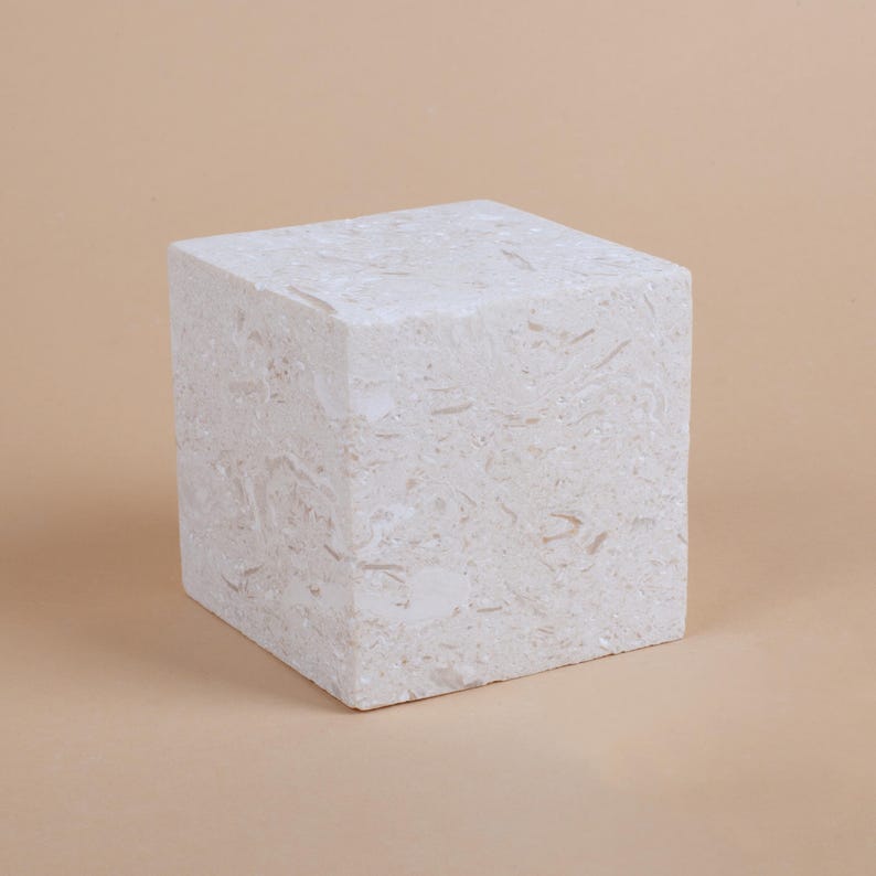 May include: A white cube-shaped pedestal made of a textured material, resembling marble or stone. The pedestal is a neutral color and has a smooth surface.