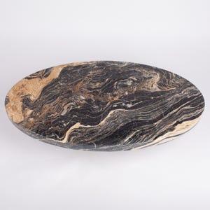 May include: Oval-shaped stone table with a polished surface. The table features a striking pattern of black, brown, and beige veining. The natural stone design adds a touch of elegance and sophistication to any space. The table measures approximately 60 inches in length.