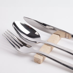 May include: A set of silver cutlery, including a fork, spoon, and knife, resting on a beige stone holder. The knife has the word "Camino" engraved on the blade.
