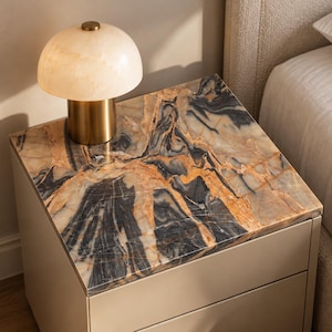 May include: A square bedside table featuring a smooth, polished stone top with intricate black, white, and amber veining. The table has a solid, light beige base with two drawers. A mushroom-shaped lamp with a translucent, cream-colored stone shade and a cylindrical brass-toned metal base sits on the surface.