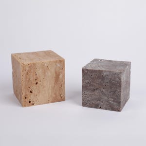 May include: Two square-shaped decorative blocks made of travertine stone. The block on the left is a light beige color with brown streaks. The block on the right is a dark gray color with white streaks.