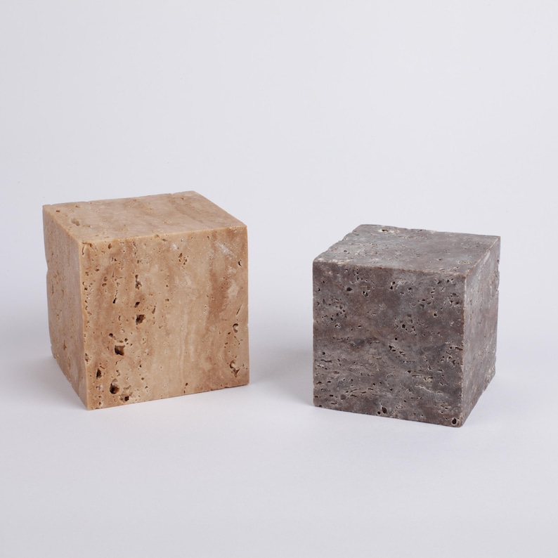 May include: Two square-shaped decorative blocks made of travertine stone. The block on the left is a light beige color with brown streaks. The block on the right is a dark gray color with white streaks.