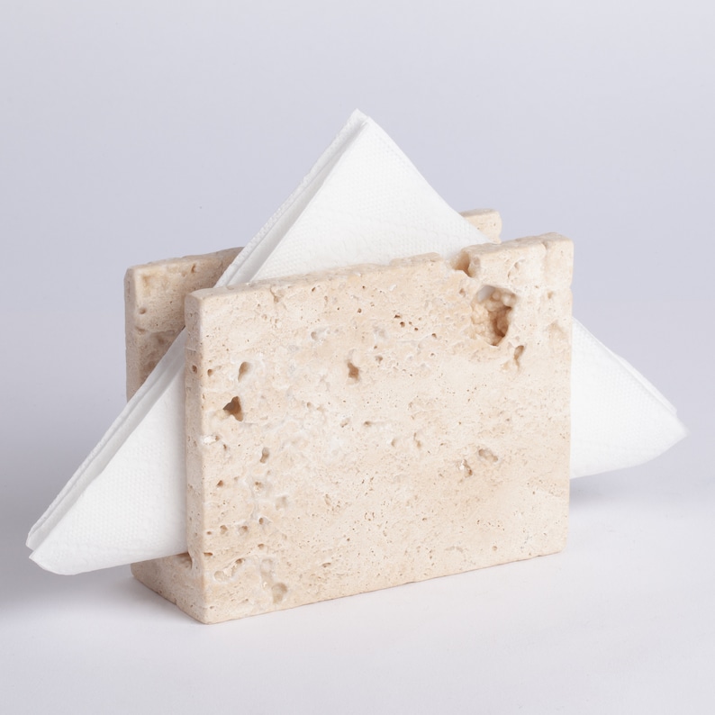 May include: A beige travertine napkin holder with a white napkin inside.