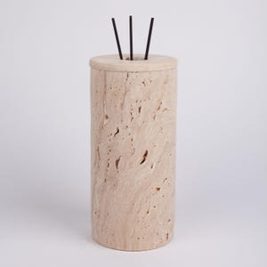 May include: A cylindrical incense holder made of light beige travertine stone with three black incense sticks sticking out of the top.