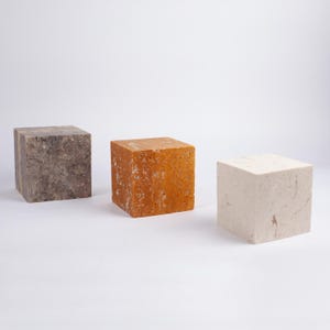 May include: Three square blocks of different colors and textures. The block on the left is gray with a rough texture. The middle block is orange with a rough texture. The block on the right is white with a smooth texture.