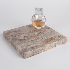 Puede incluir: Un frasco de perfume transparente y facetado con tapón plateado, lleno de líquido ámbar, se encuentra sobre una losa de piedra rectangular texturizada. La piedra presenta un patrón moteado de tonos grises, marrones y beige.