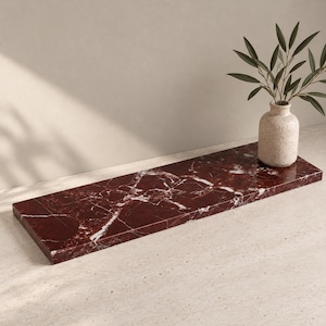 May include: A rectangular floating shelf made of deep red marble with prominent white veining. A light-colored, textured ceramic vase containing a green leafy plant sits on the right side of the shelf, which is positioned horizontally.