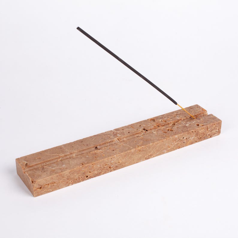 Pode incluir: Um porta-incenso retangular de m&aacute;rmore castanho com duas ranhuras paralelas. Um pau de incenso preto com ponta dourada &eacute; inserido no suporte, inclinado para cima. O suporte est&aacute; sobre um fundo branco.