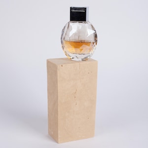 May include: A clear glass bottle with a silver cap and a light brown liquid inside, sitting on a beige rectangular pedestal.