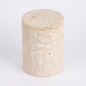 May include: A cylindrical beige stone container with a lid. The container has a rough, textured surface with white and brown markings.