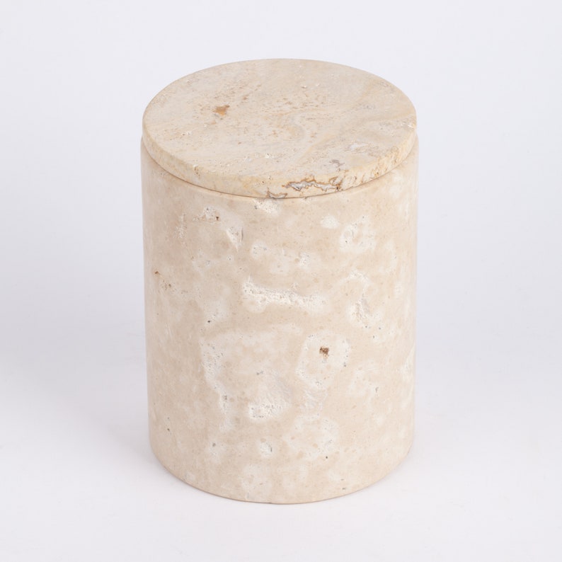 May include: A cylindrical beige stone container with a lid. The container has a rough, textured surface with white and brown markings.