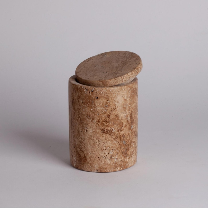 May include: A cylindrical stone container with a lid. The container is made of travertine and has a natural brown and beige color with a marbled pattern.