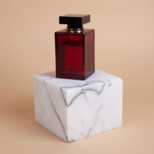May include: A red glass perfume bottle with a black cap sits on a white marble pedestal. The bottle is filled with a red liquid.