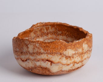 Puede incluir: Un cuenco de piedra rayado en marrón y blanco con una textura rugosa e irregular. El cuenco está hecho de piedra natural y tiene una forma única y orgánica.