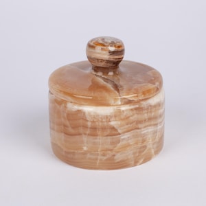 May include: A small, round, lidded jar made of layered, tan-coloured stone. The jar has a cylindrical body and a rounded lid with a small, knob-like handle. The stone has a natural, marbled appearance with variations in colour.