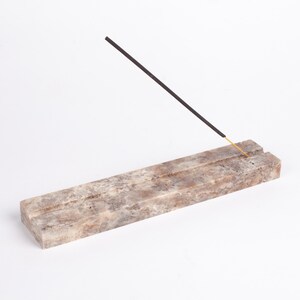Pode incluir: Um porta-incenso retangular feito de pedra bege e castanha. Um incenso preto com ponta dourada &eacute; inserido no suporte, inclinado para cima. O suporte tem uma superf&iacute;cie texturizada e uma ranhura para segurar o incenso.
