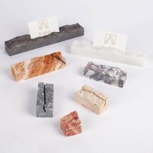 Puede incluir: Una colección de tarjeteros de piedra en varios colores y texturas. Los soportes son rectangulares con una ranura para sujetar una tarjeta. Algunos soportes tienen un aspecto rugoso y natural, mientras que otros son lisos. Un soporte muestra una tarjeta con el nombre "Andrés" y un logotipo.