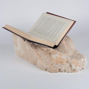 May include: An open hardcover book with cream-colored pages and dark brown covers resting on a large, rough-hewn piece of beige natural stone. The stone has a porous, textured surface with irregular edges, serving as a sturdy base for the open book.