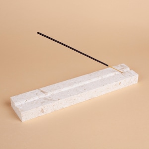 Pode incluir: Um porta-incenso retangular branco sujo com uma superf&iacute;cie texturizada. O suporte tem duas ranhuras paralelas e um &uacute;nico pau de incenso &eacute; colocado em uma das ranhuras. O pau de incenso &eacute; preto e tem uma ponta dourada.