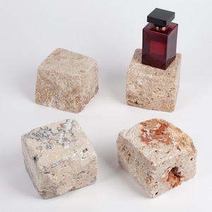 May include: Four rough-hewn, cubic stone blocks with porous, natural textures in shades of beige, tan, and reddish-brown. One block serves as a base for a rectangular, deep red glass perfume bottle with a black cap, displaying a clear liquid inside.