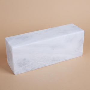 May include: A rectangular block of white marble, featuring delicate grey veining. The stone has a polished surface and a minimalist design. The block is a solid, rectangular shape, suitable for decorative or functional use.
