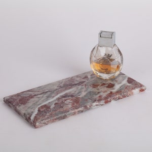 May include: A rectangular marble tray with a pink, gray, and white marbled pattern. A clear glass bottle of perfume with a silver cap sits on the tray.