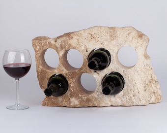 Puede incluir: Un estante de vino de piedra con forma de roca con seis ranuras para botellas. El estante está hecho de piedra marrón claro y tiene una textura rugosa y natural. Hay cuatro botellas de vino en el estante.