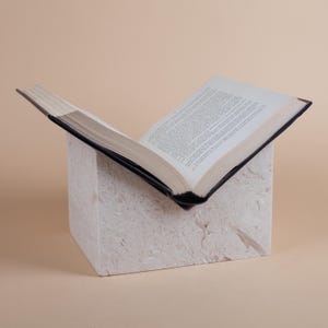 Könnte beinhalten: Ein offenes Buch mit weißen Seiten und einem schwarzen Einband liegt auf einem weißen, strukturierten Block. Das Buch ist auf einer Seite mit viel Text aufgeschlagen.