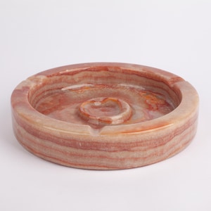 Puede incluir: Un cenicero redondo de mármol rosa y marrón con una pequeña hendidura redonda en el centro.