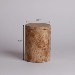 May include: A cylindrical, beige travertine stone container with a lid. The container measures 5.1 inches tall and 3.7 inches in diameter.
