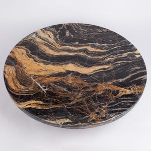 Op de afbeelding: Een ronde marmeren tafelblad in zwart met gouden en witte adering. De gepolijste steen heeft een natuurlijk patroon. Geschikt voor binnenshuis, de tafel voegt een luxe uitstraling toe. Diameter ongeveer 76 cm.