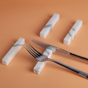 Pode incluir: Um garfo e uma faca de aço inoxidável repousam sobre uma superfície de mármore branco com veios cinzentos. A faca tem a palavra "Camino" gravada na lâmina. Quatro descansos de mármore adicionais são dispostos ao redor dos utensílios sobre um fundo laranja.